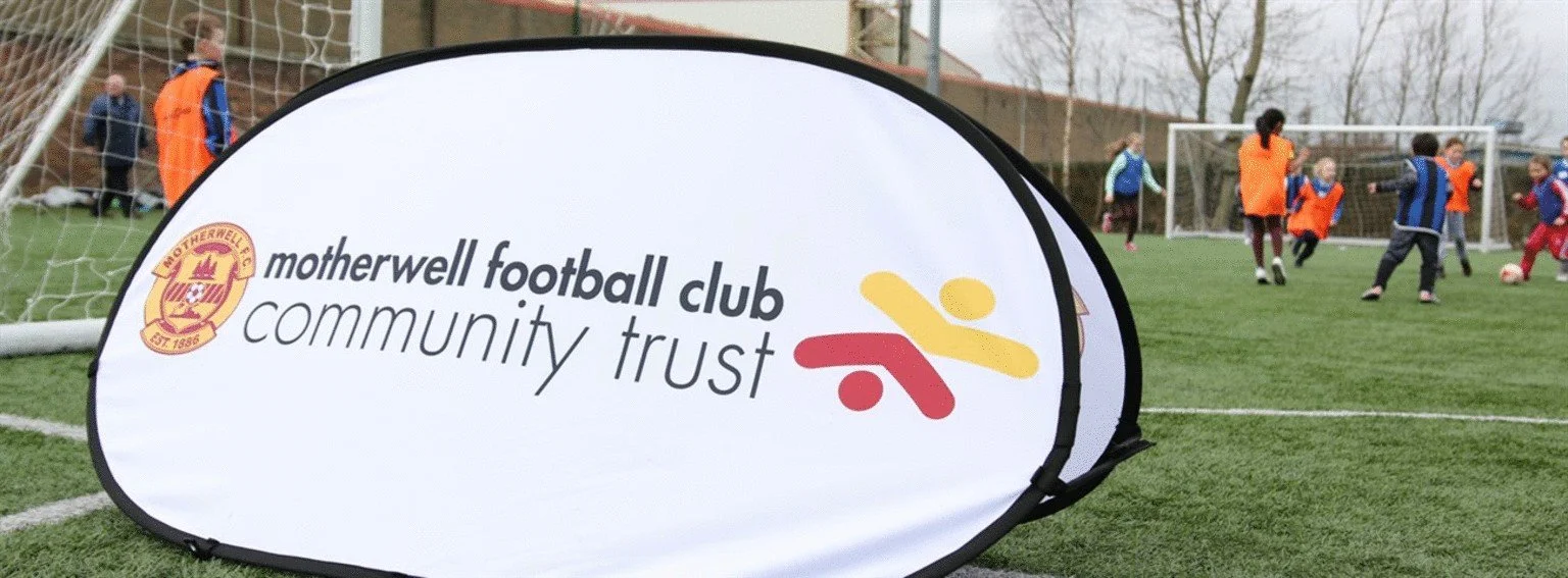 Soccer field with children playing on it, a large white sign with the Motherwell Football Club Community Trust logo and name, and people in the background.