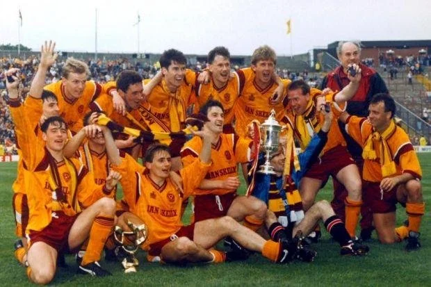 Soccer team celebrating victory on field, holding a trophy and medals, with spectators in the background.