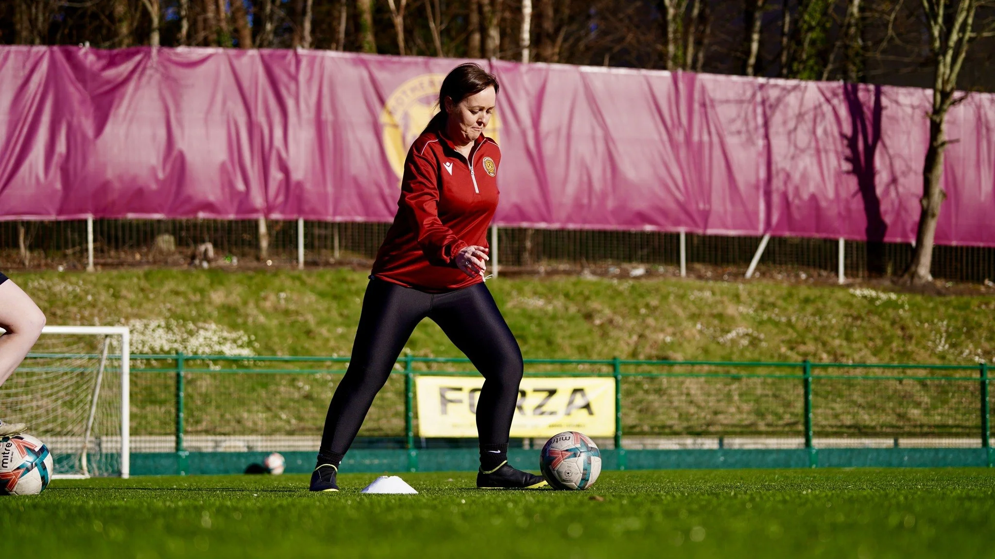 A woman in a red sports jacket practicing soccer on a field with a pink barrier in the background.