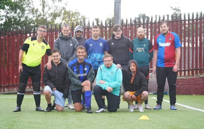 Group of ten young men on a soccer field, some kneeling and others standing, dressed in sportswear and casual clothing, with a red fence and trees in the background.