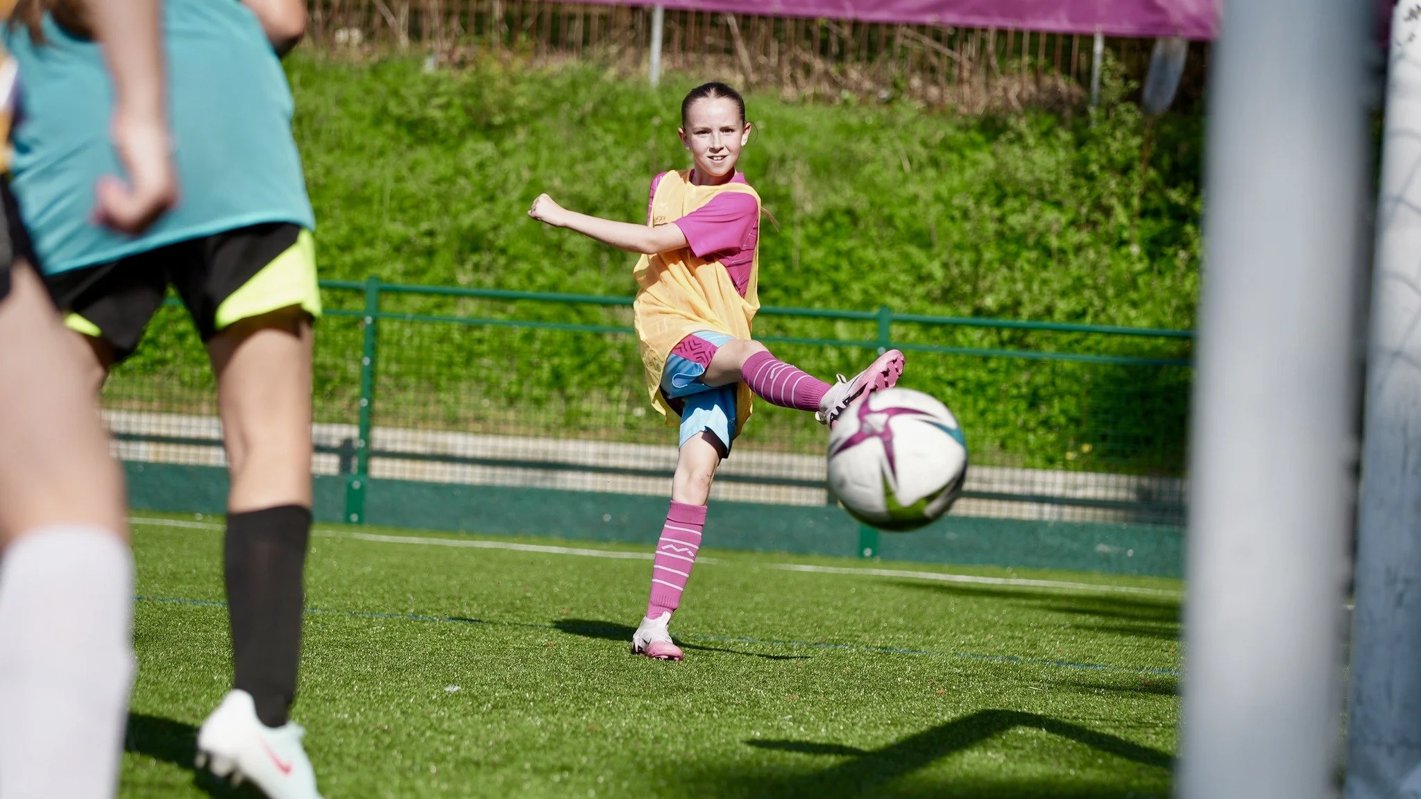 A young girl in pink and yellow sportswear kicking a football on a green field during a game.