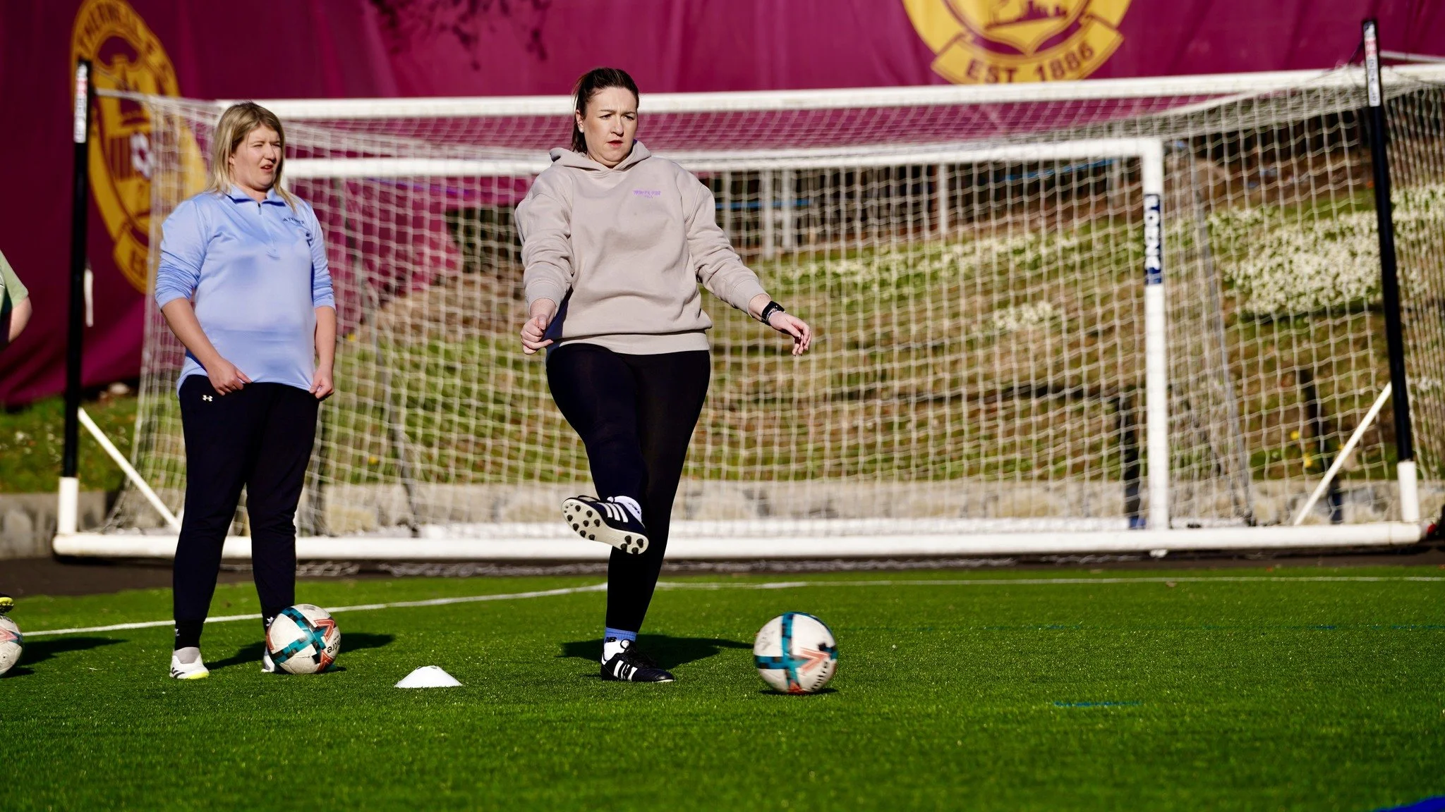 Women practicing soccer drills on a field, one woman is balancing a soccer ball on her foot, with a goal net in the background.