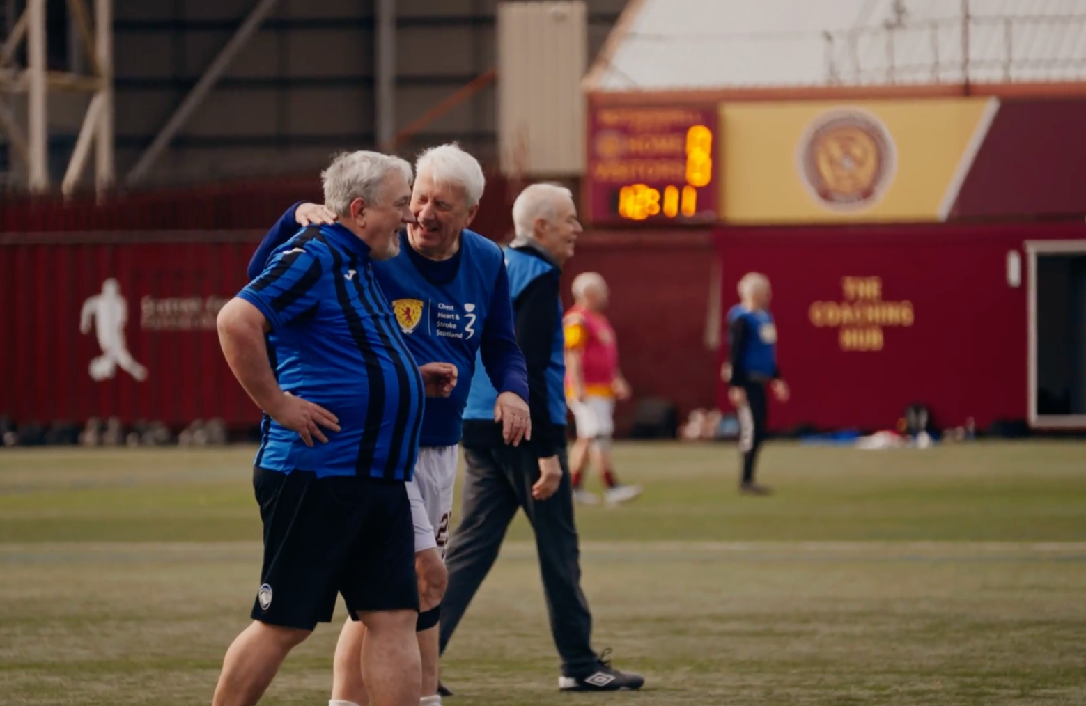 Older men playing or celebrating on a soccer field inside a stadium, wearing blue jerseys, with a scoreboard and red seating in the background.