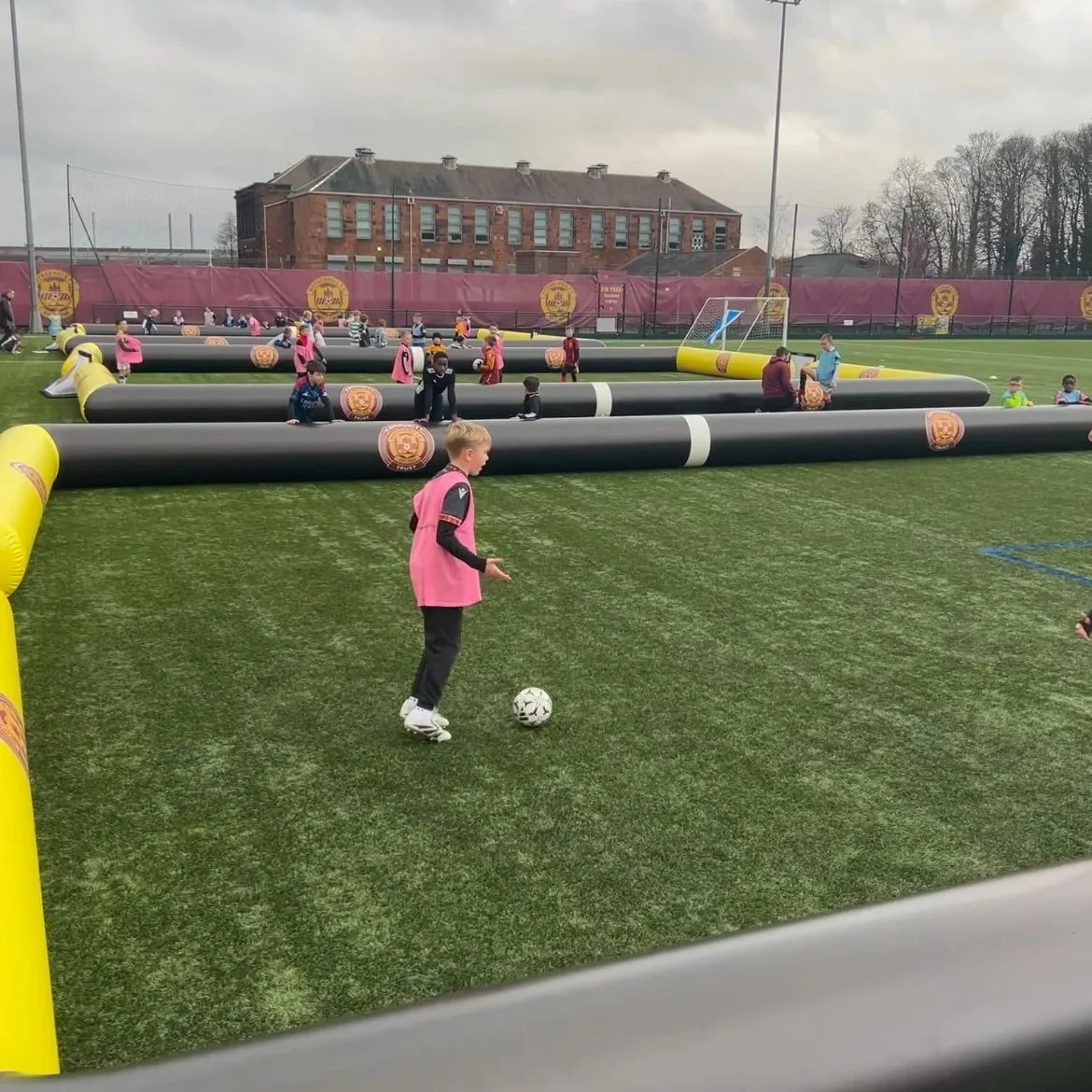 Children practicing football on a field with inflatable barriers and a soccer goal, in a sports complex with a building and trees in the background.