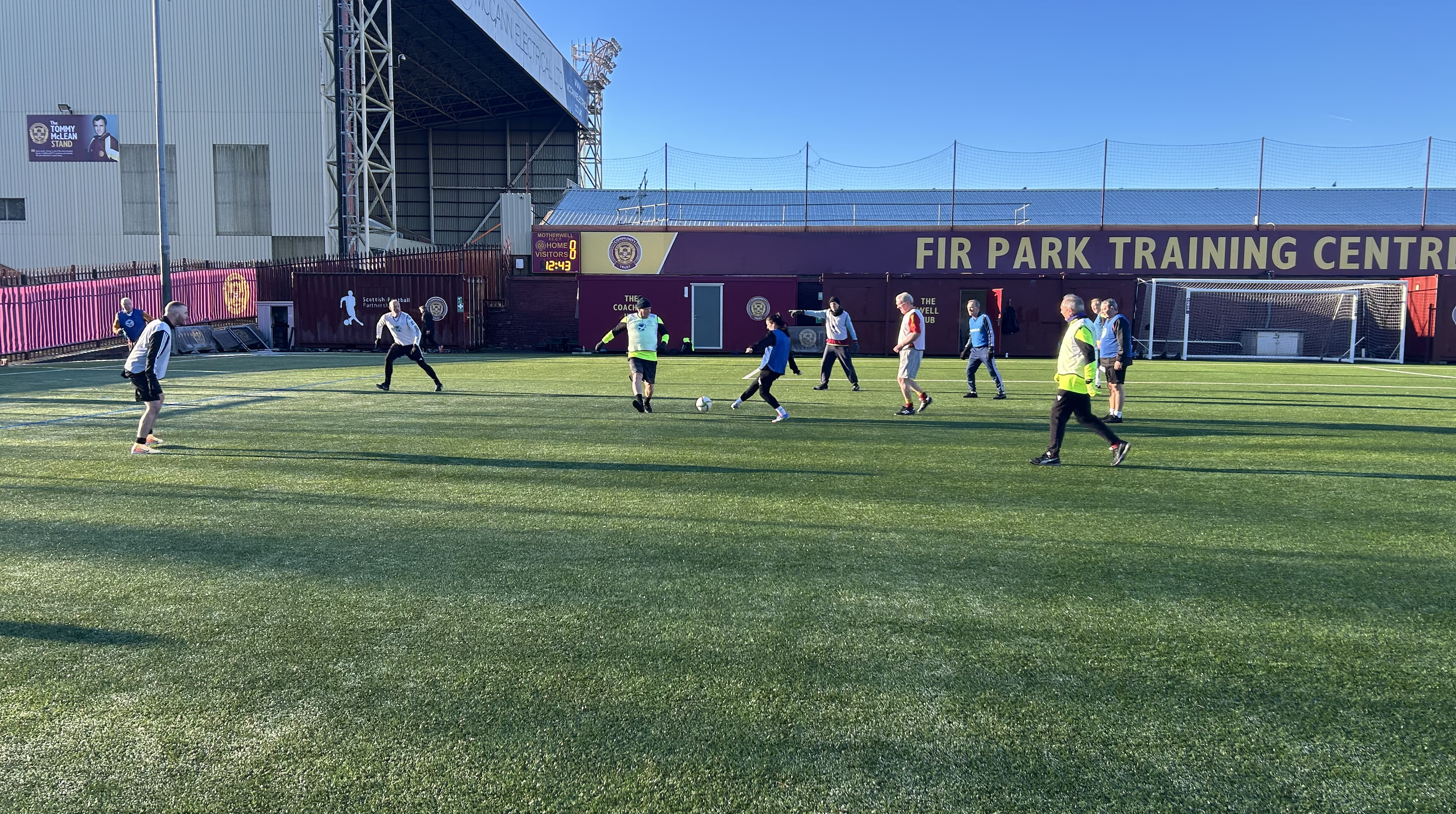 A group of people playing soccer on a field at Fir Park Training Center, with a stadium and scoreboard in the background.