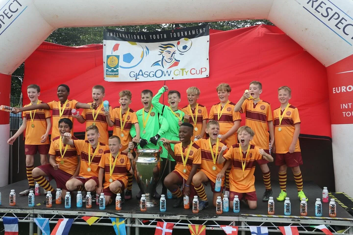 Young boys in MFCCT strips celebrating on a podium after winning a tournament, with medals around their necks and water bottles in hand, under an inflatable arch with the Glasgow City Cup logo.