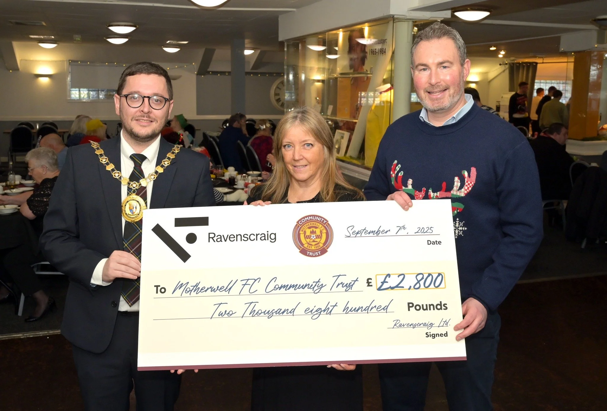 Three people at an event holding a large check. The person on the left is a man wearing glasses, a suit, and a chain of office. The woman in the middle has blonde hair, and the man on the right has short gray hair and a beard, wearing a festive sweater. The check is made out to Motherwell FC Community Trust for £2,800, dated September 7th, 2025, and signed by Ravenscraig Ltd.