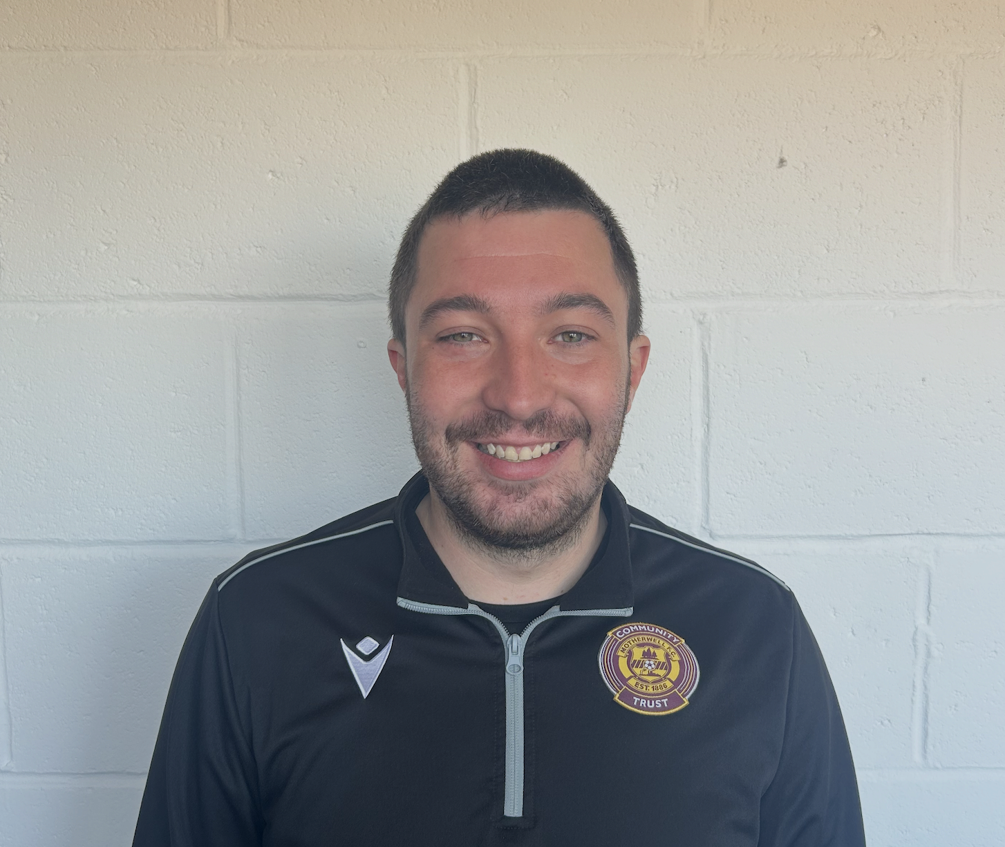 A man smiling, wearing a black sports jacket with a Johnstone's Community Trust emblem on the chest, standing against a light-colored brick wall.