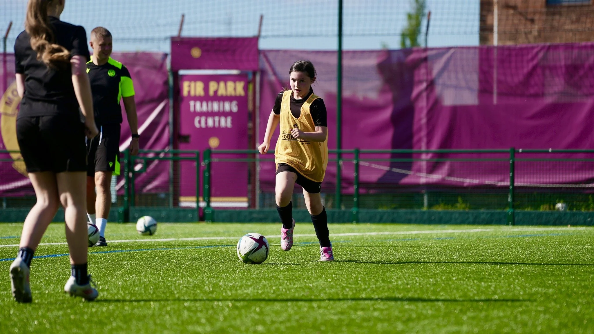 Young girl in a yellow training vest kicking a soccer ball on a field, with others and a goal in the background.