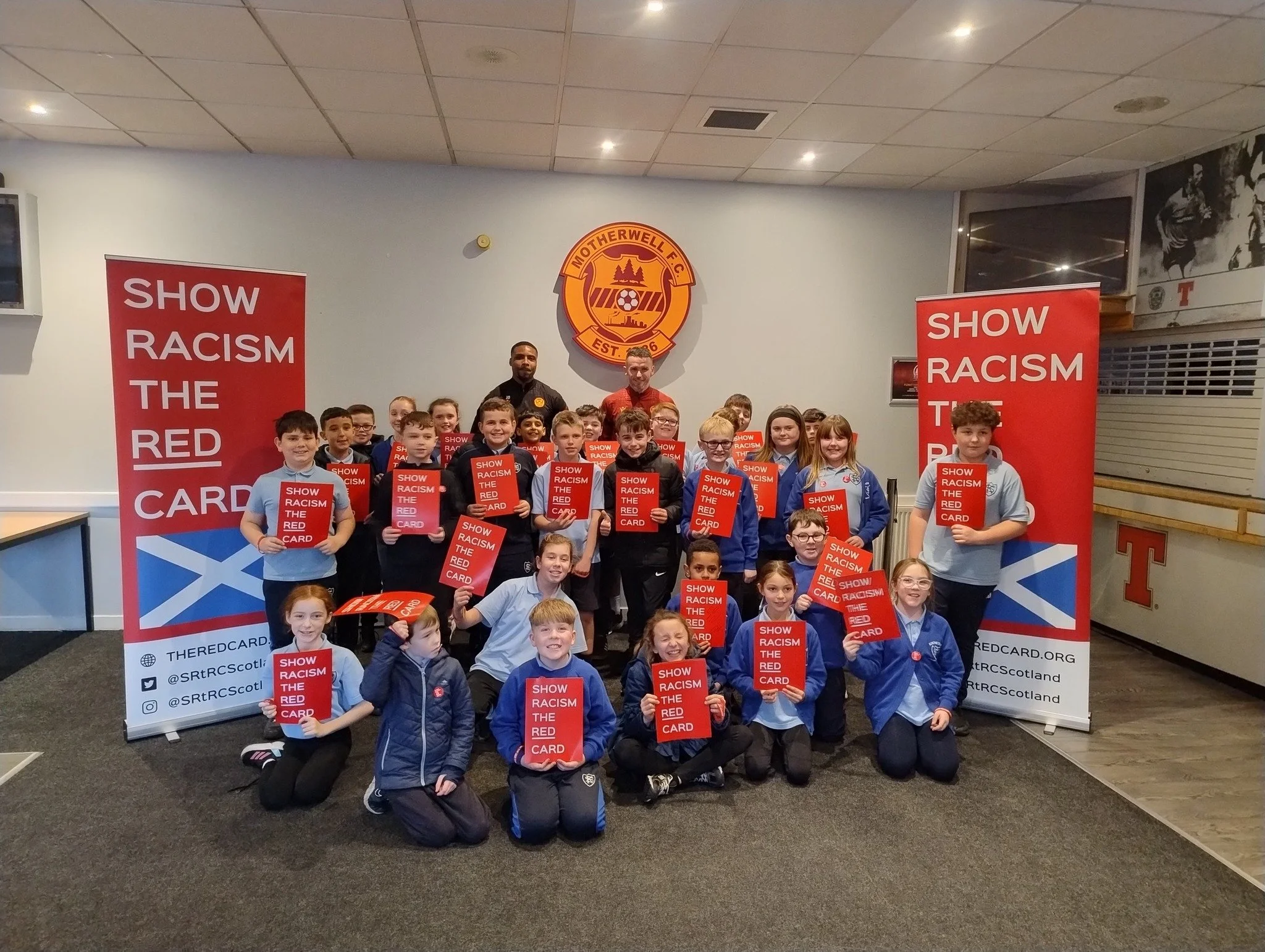 Group of children and adults holding red signs that read "Show Racism the Red Card" in front of a Motherwell F.C. emblem and banners supporting anti-racism.