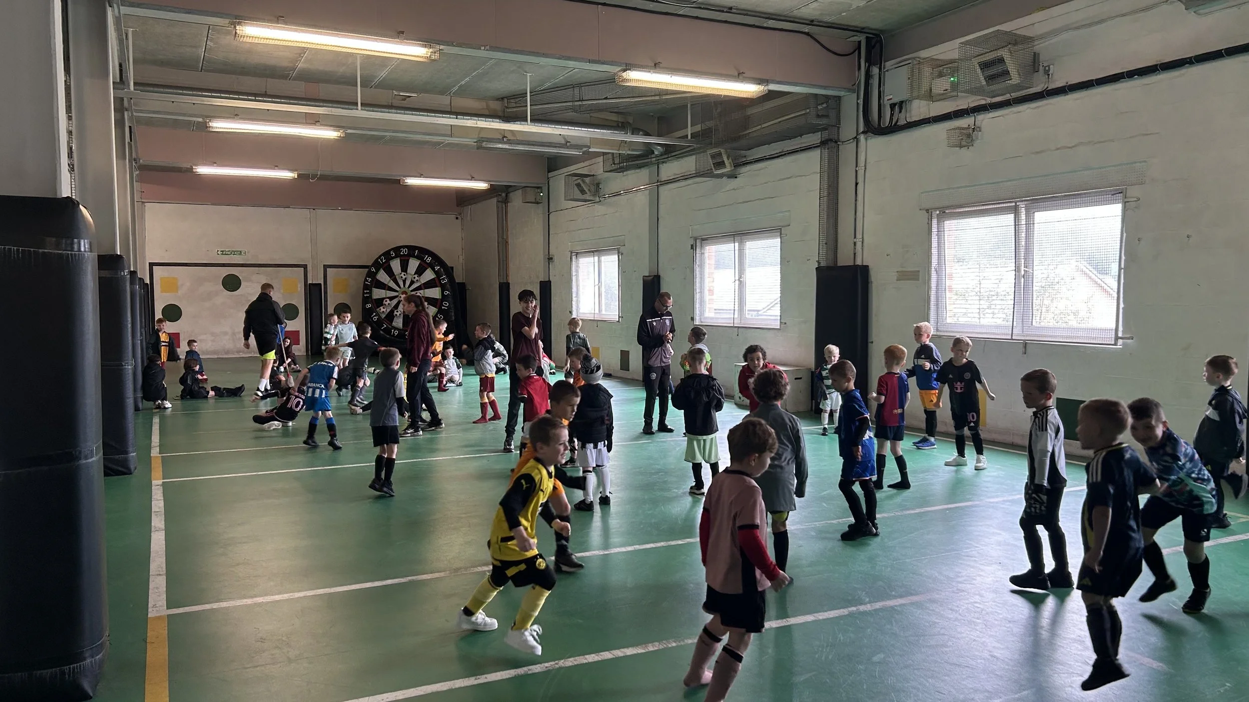 Children participating in a sports activity on a green indoor court, with some adults supervising, inside a gymnasium with large windows and a dartboard on the wall.