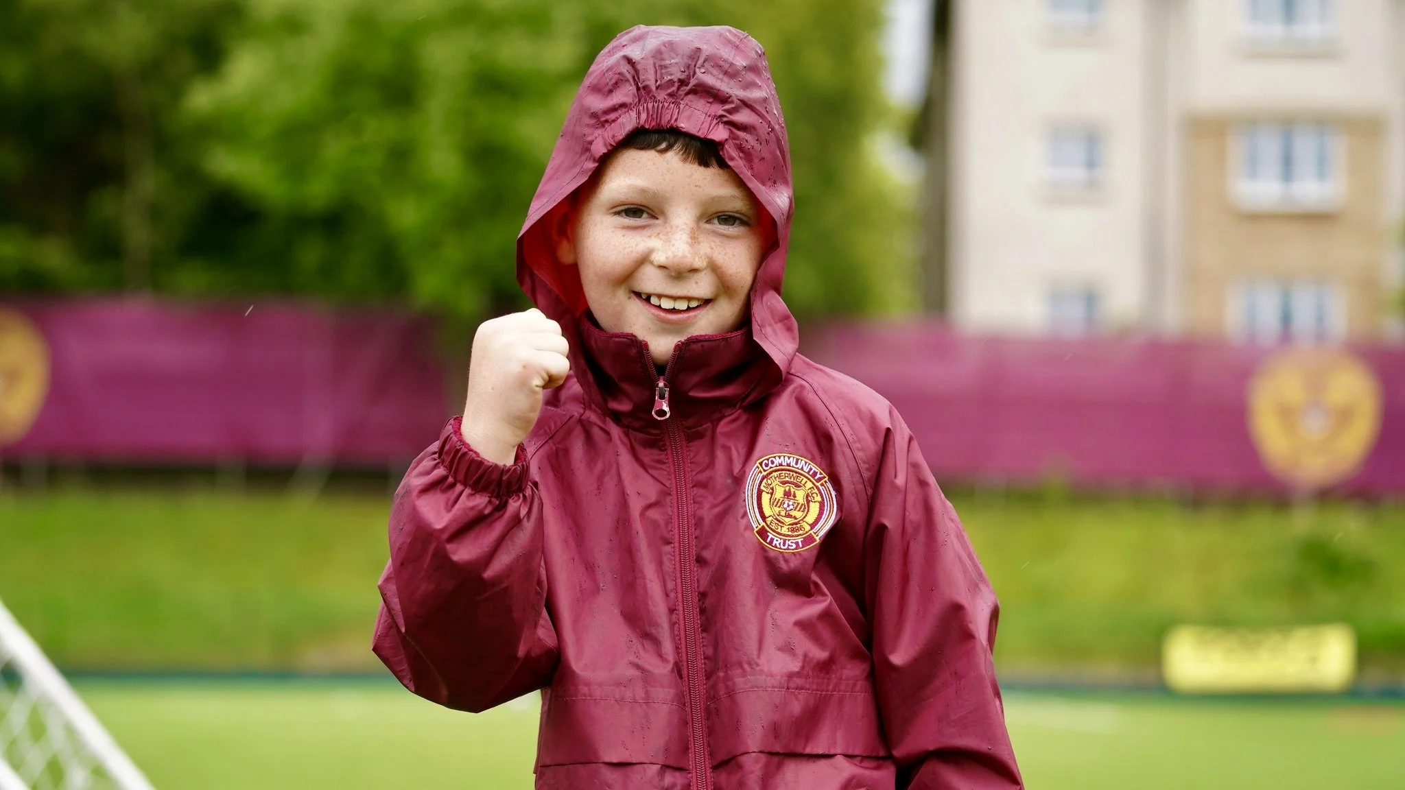 A young boy wearing a claret rain jacket with a hood, smiling and making a fist gesture outdoors with green trees and buildings in the background.