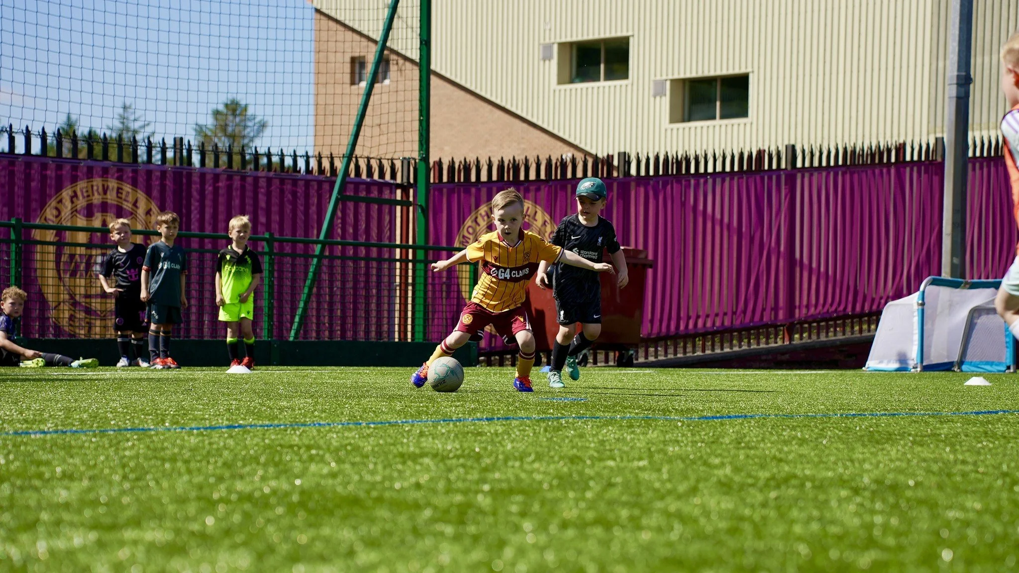 Children playing soccer on a green field with a purple fence and a yellow building in the background.