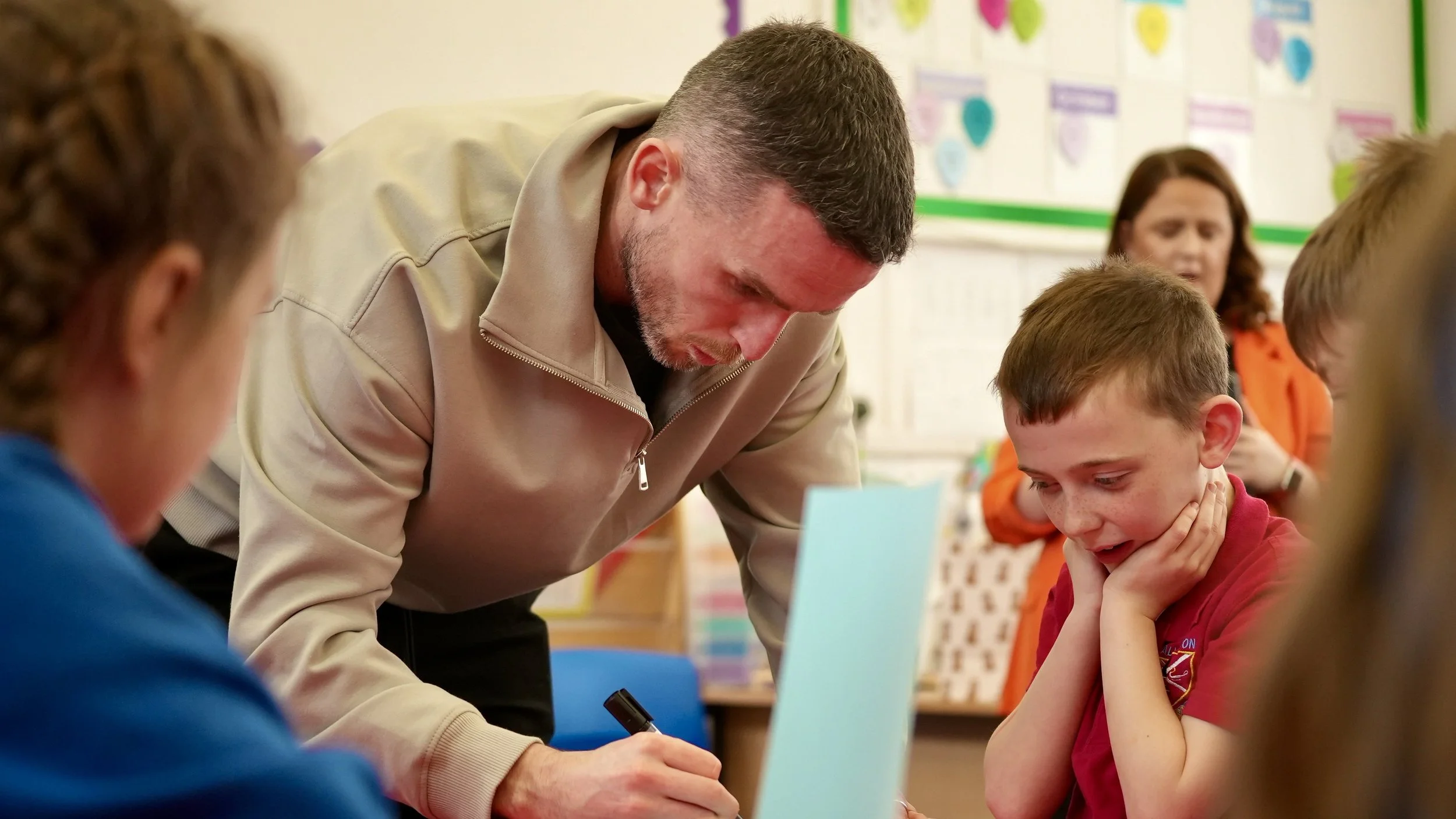 A teacher or adult leaning over a student in a classroom, helping with their work. The student, wearing a red shirt, is holding his chin and looking down at a laptop or paper. Other children are visible in the background along with educational posters on the wall.