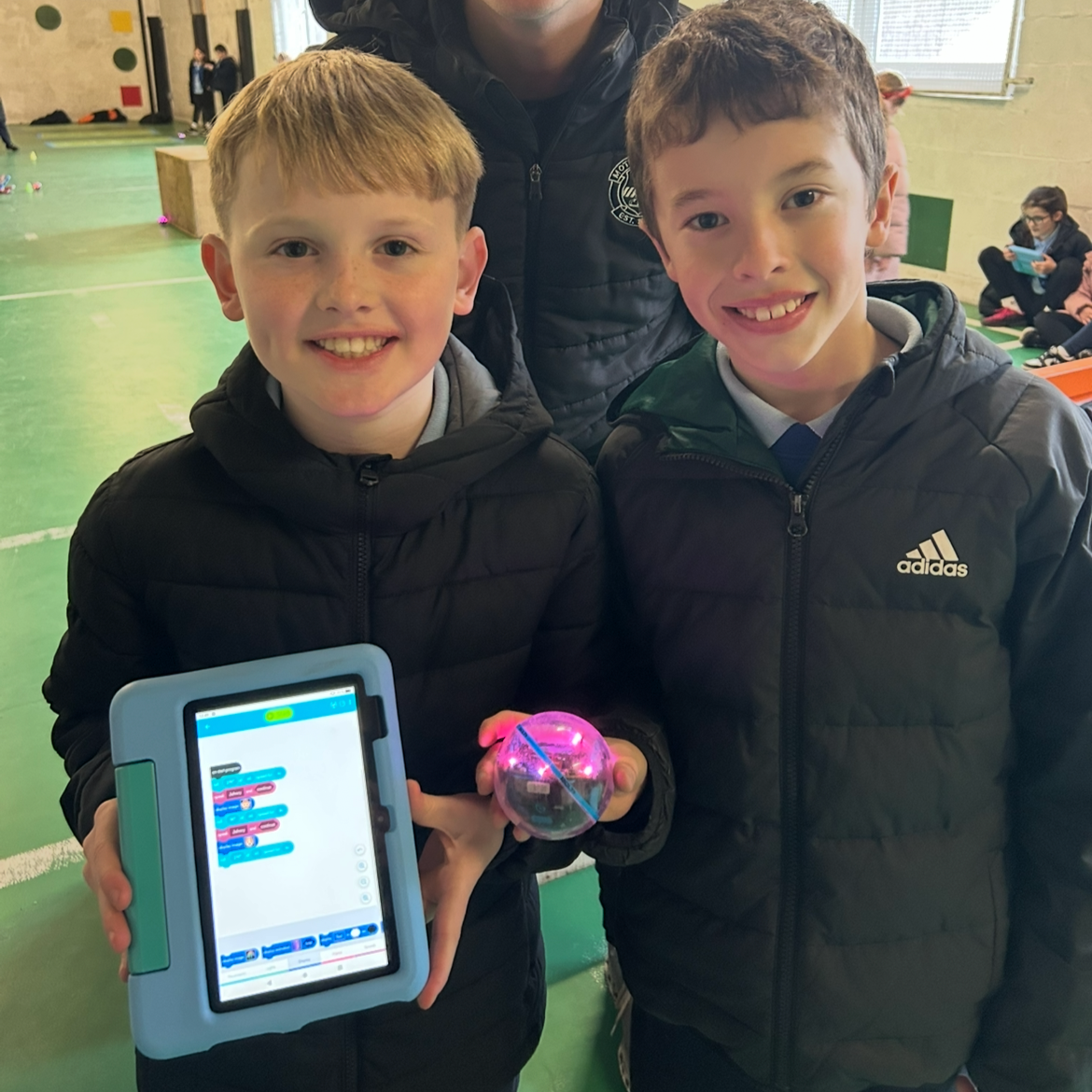 Two boys smiling and holding a small electronic device and a glowing spherical object indoors, with other children seated in the background.