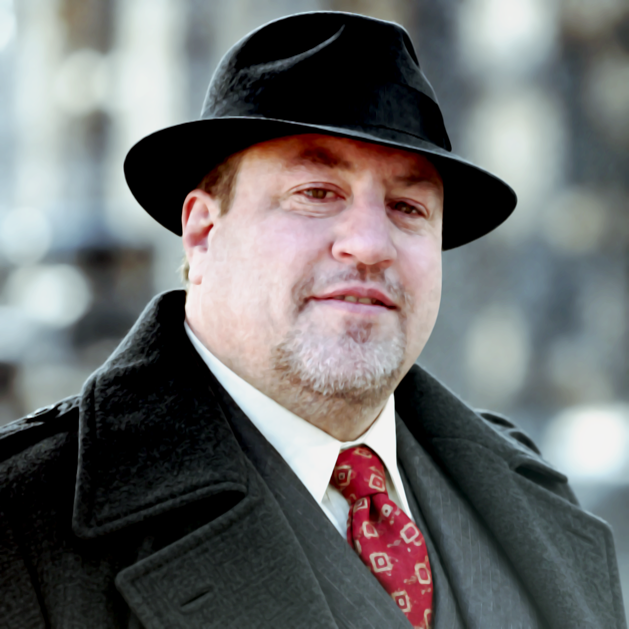 Brian J. Eskridge wearing a black fedora hat, a dark coat, a white shirt, and a red patterned tie, standing outdoors with blurred background.