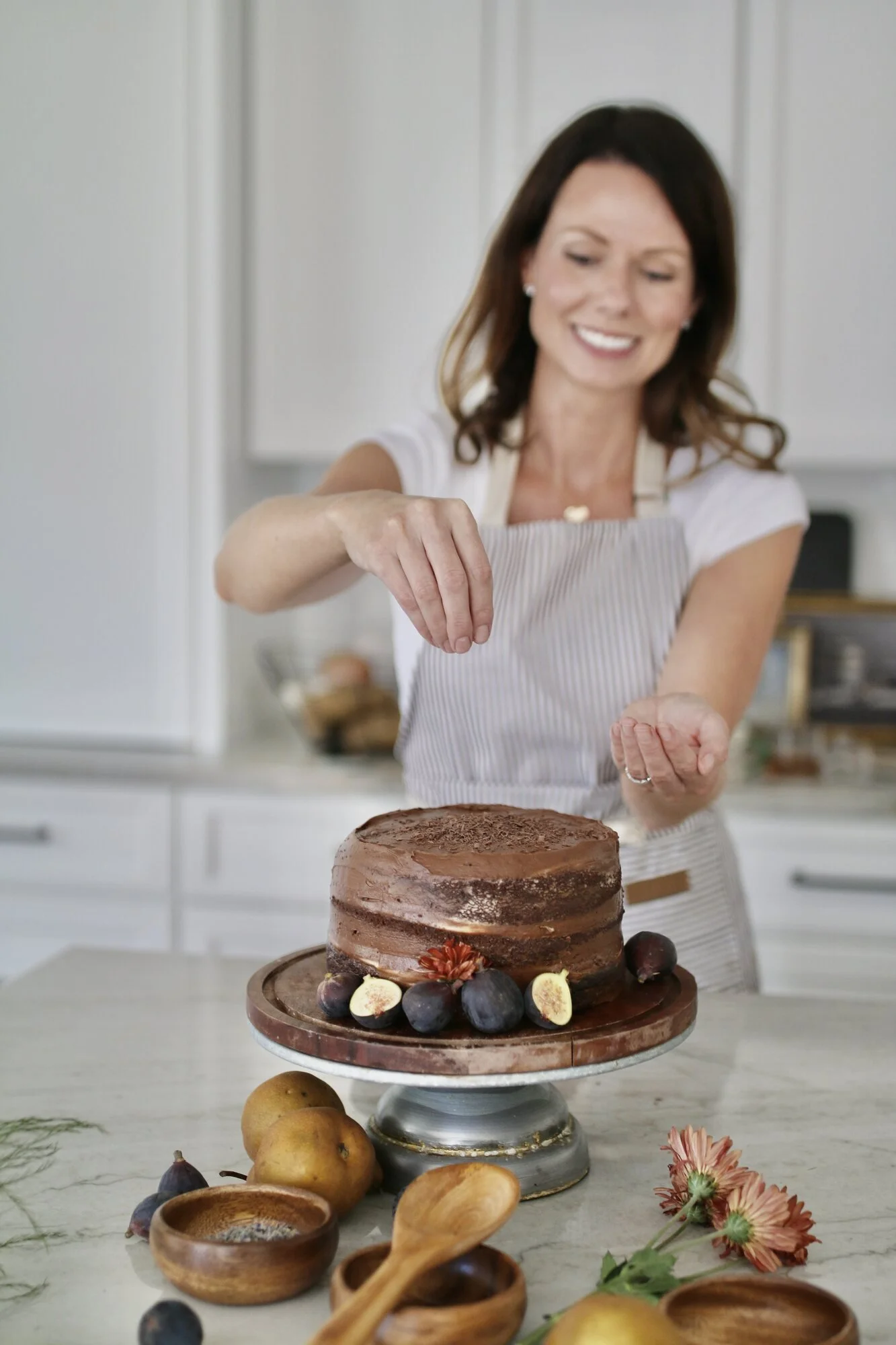 Mary Beth Maly sprinkling shaved chocolate over a homemade chocolate cake in her kitchen while wearing an apron.