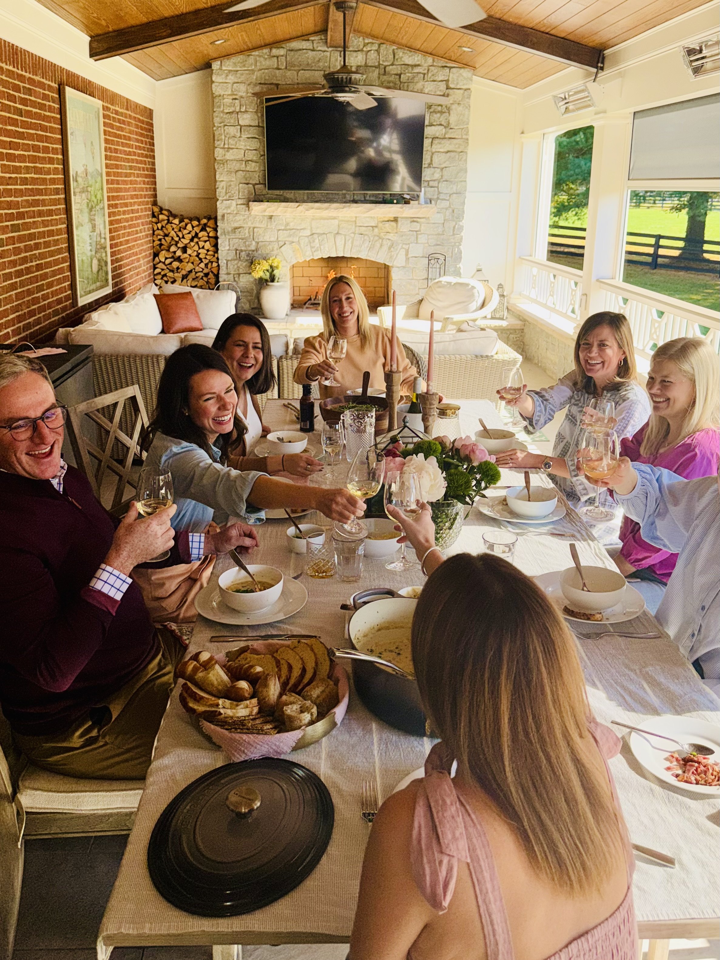 Seven guests gathered around an outdoor table by a fireplace, toasting with white wine over a summer meal with flowers and centerpieces.