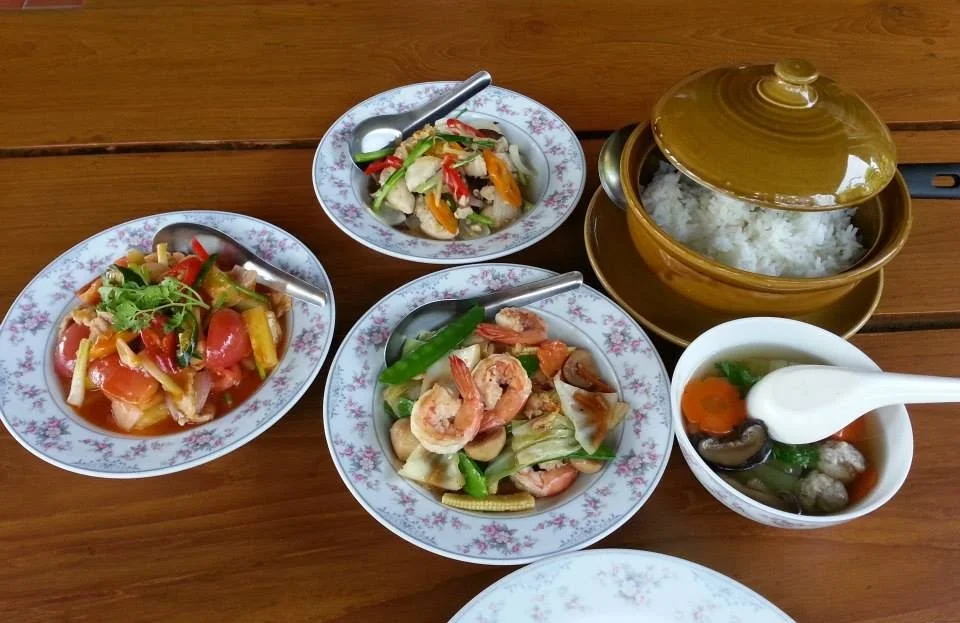 Assorted Asian dishes on a wooden table, including rice in a covered brown pot, stir-fried vegetables with shrimp, tomato salad, and a bowl of soup with vegetables and meatballs.