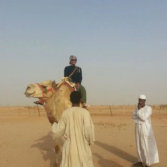 A woman riding a camel in a desert, flanked by two men dressed in traditional white robes and head coverings.