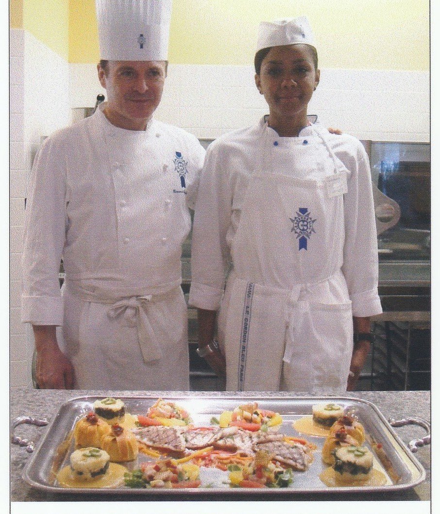 Two chefs in white uniforms and tall hats standing behind a tray of plated food in a kitchen.