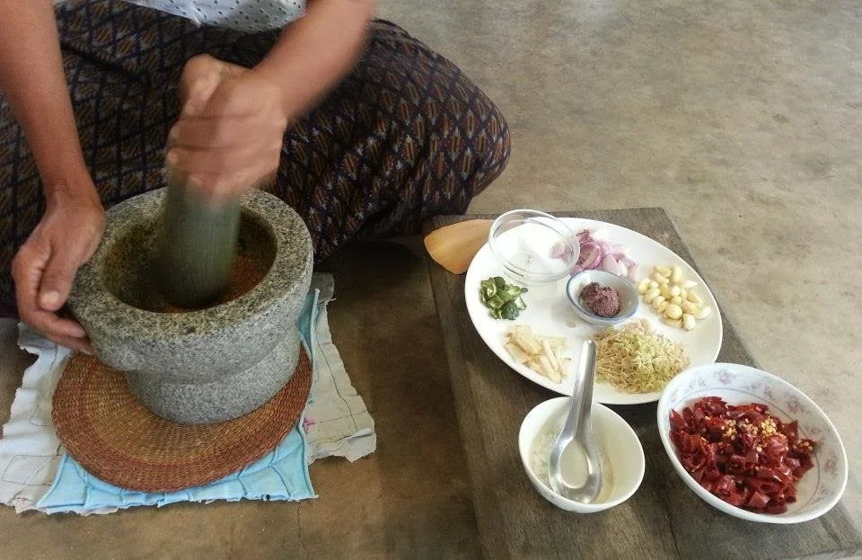 Person using a pestle to grind ingredients in a stone grinder. Various ingredients on a white plate and bowls, including garlic, shallots, chili peppers, and herbs, on a wooden table.
