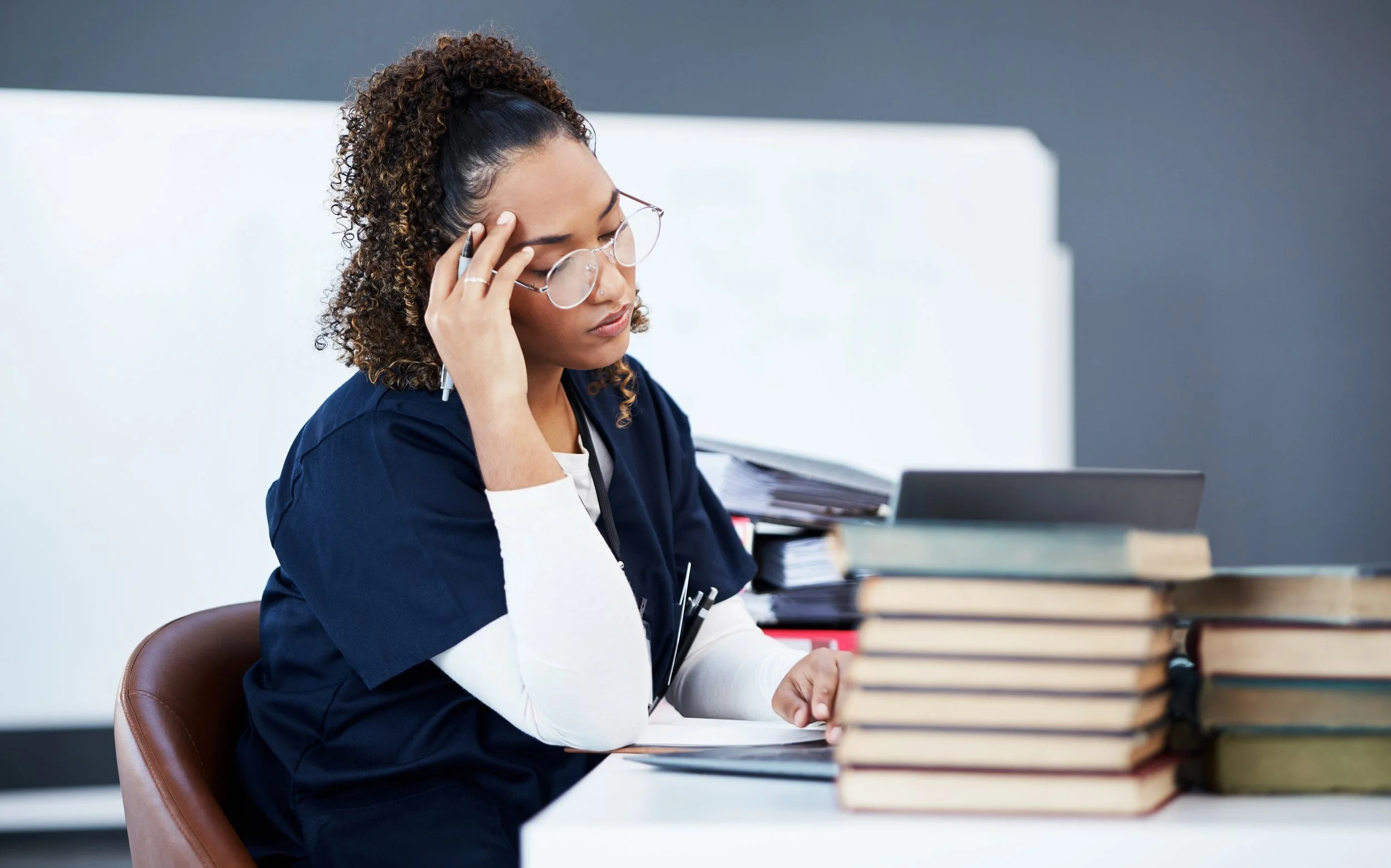 A woman with curly hair tied in a high bun, wearing glasses and a navy blue medical scrub, sits at a desk surrounded by stacks of paper and books, resting her head on her hand with a tired or stressed expression.