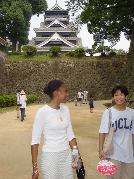 Two women walking outdoors near a historic Japanese castle with trees and other visitors in the background.