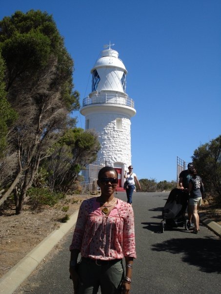 Woman in pink blouse standing on pathway near a lighthouse with other visitors and trees in the background.