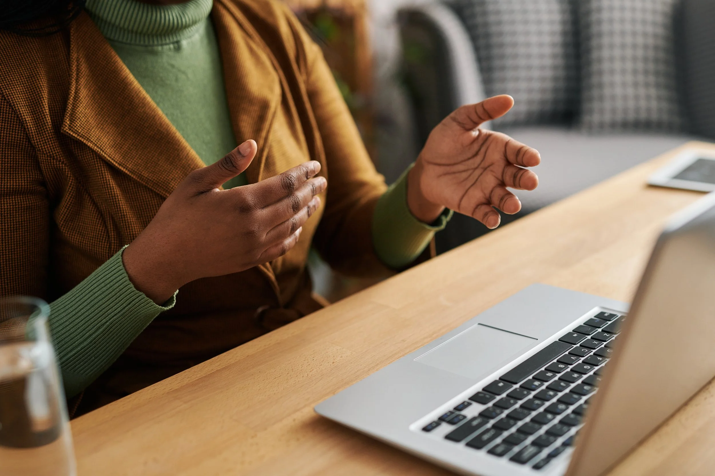 Close-up of a person wearing green sweater and brown jacket, gesturing with hands near a laptop on a wooden table, in a meeting or discussion setting.