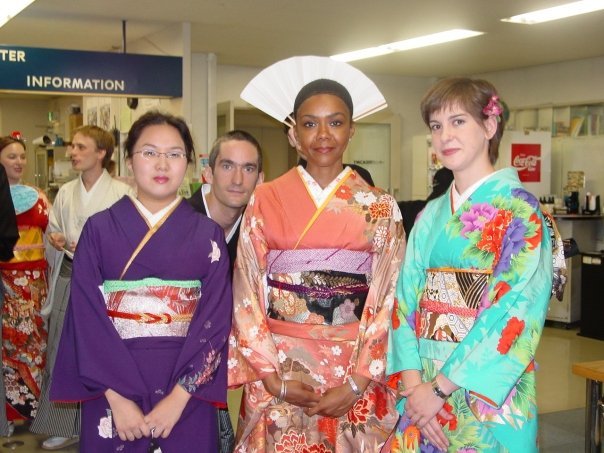 Group of four people in traditional Japanese kimonos and a woman in Western clothing in an indoor setting, possibly an airport or train station.