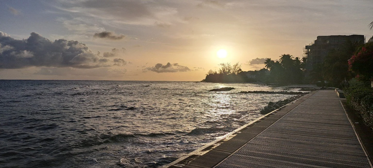 Sunset over the ocean with a wooden walkway along the beach, trees, and buildings on the right side.