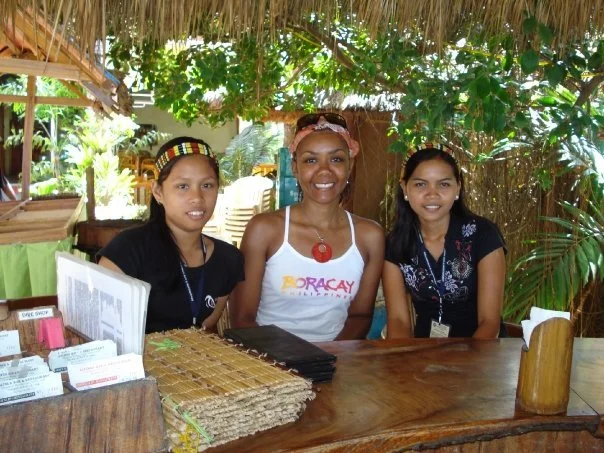 Three women sitting at a wooden table outdoors, surrounded by green plants and trees, under a thatched roof. They are smiling and looking at the camera.
