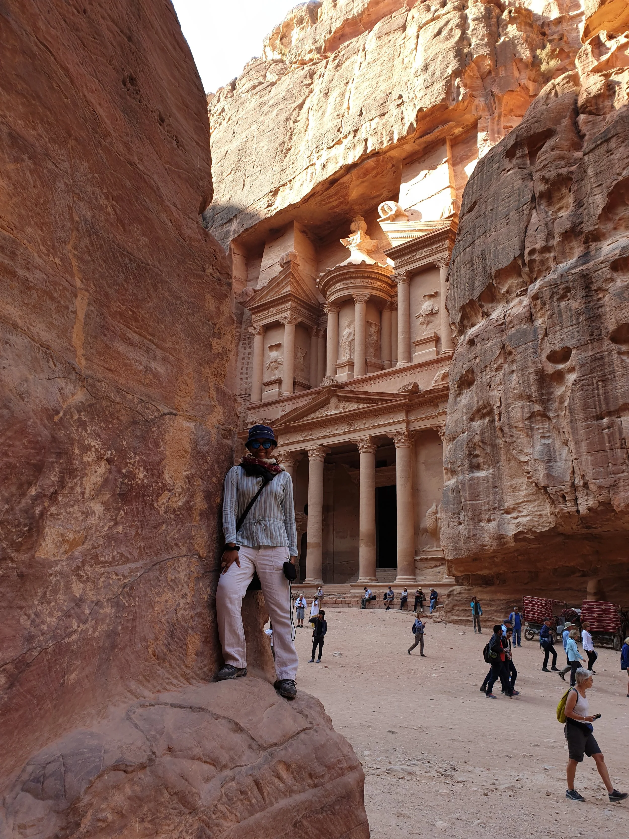 A person standing in front of the ancient city of Petra, carved into pink sandstone cliffs in Jordan, with tourists walking around and the iconic Treasury building visible in the background.