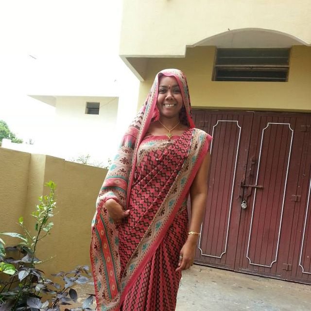 A smiling woman dressed in a pink and beige traditional saree standing outdoors in front of a building with a wooden door and a small window, with plants nearby.