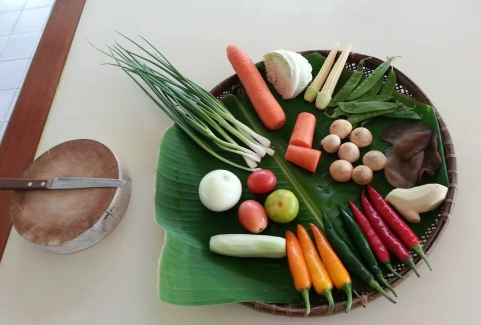 Assorted fresh vegetables including green onions, a carrot, garlic, spring onions, tomatoes, apples, mushrooms, chili peppers, and leafy greens on a green banana leaf, with a knife on a wooden chopping board nearby.