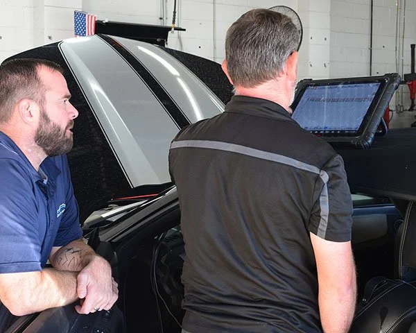 Two men working on a car, one with a beard leaning on the car and another with gray hair using a tablet or diagnostic tool, inside a garage.