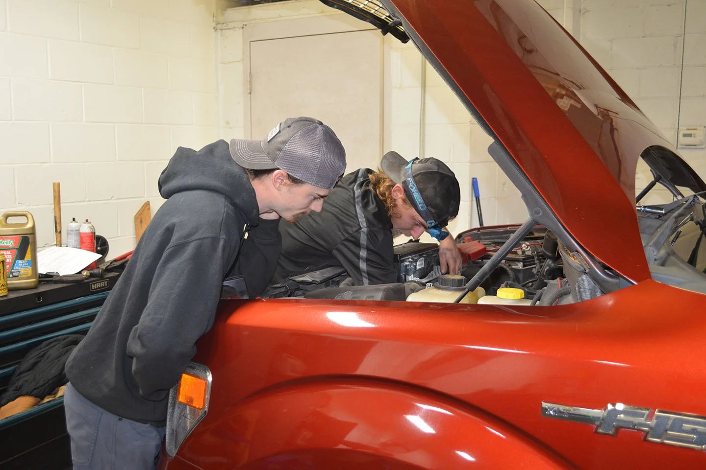 Two men working on a red Chevrolet vehicle with its hood open in a garage.