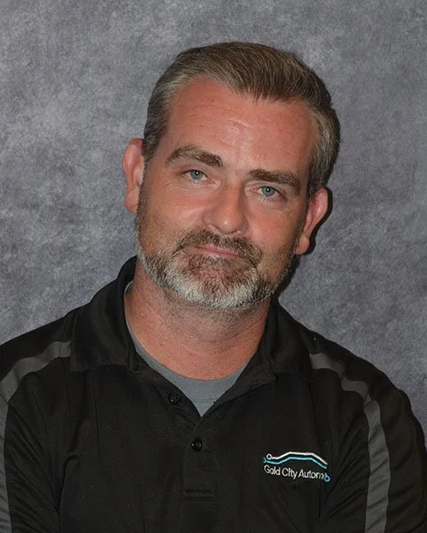 A man with gray hair and beard wearing a black collared shirt with a logo, standing against a gray background.