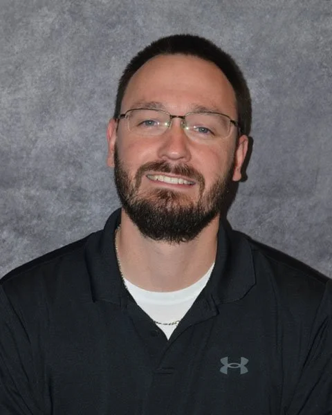 Portrait of a smiling man with glasses, short dark hair, and a beard, wearing a black Under Armour polo shirt, against a gray textured background.