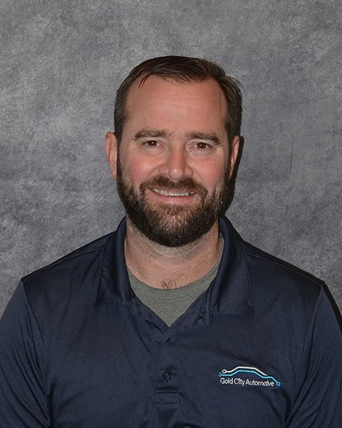 A man with brown hair, a beard, and a mustache smiling, wearing a navy blue polo shirt with a logo on the left chest that says 'Gold City Automotive' against a gray background.