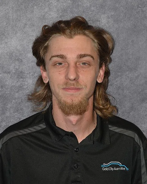 A young man with dark hair, a goatee, and a mustache, wearing a black polo shirt with a logo that says 'Gold City Automotive,' standing against a gray background.
