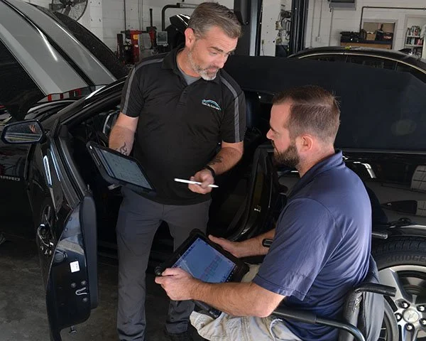Two men, one standing and one seated in a wheelchair, discussing car information using tablets inside a garage or service center.