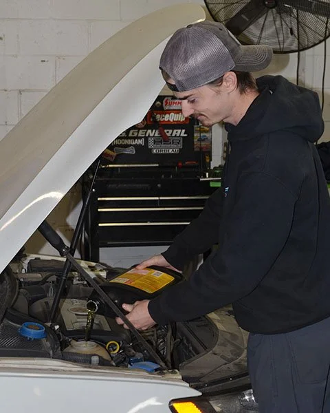 Young man in a gray cap and black hoodie checking the car's engine with the hood up in a garage.
