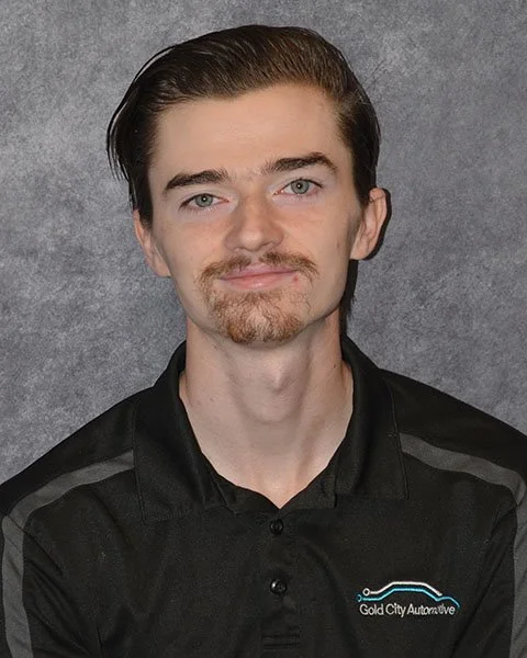 A young man with long, wavy hair, light facial hair, and a slight smile, wearing a black collared shirt with 'Gold City Automotive' logo, standing against a gray background.