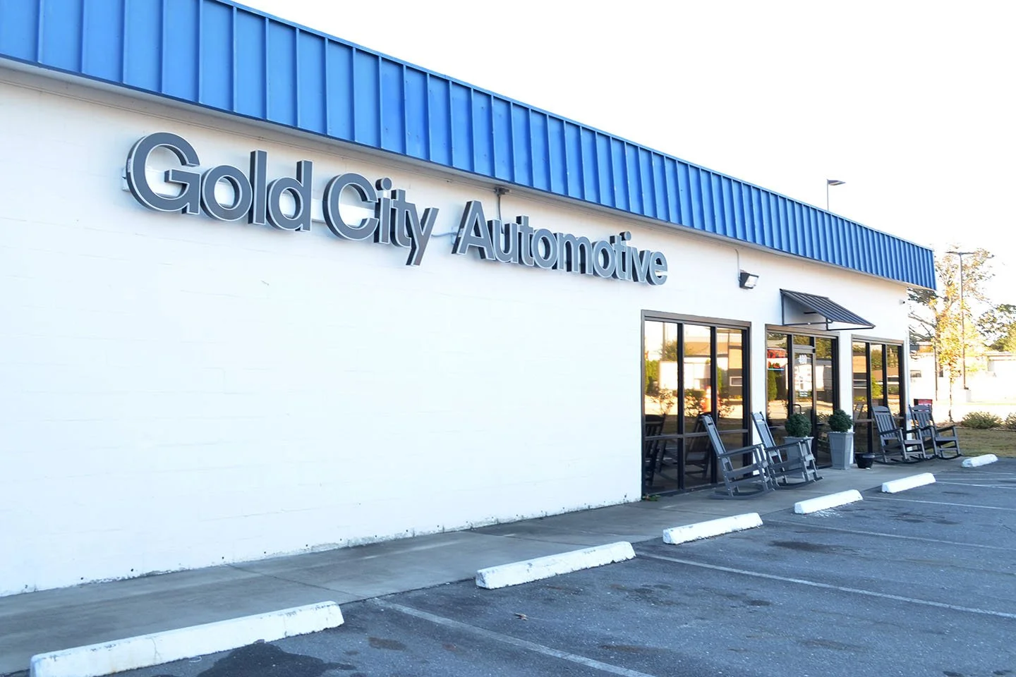 Exterior of a building with a sign that reads 'Gold City Automotive,' featuring a white brick wall, large windows, a blue metal roof, and a parking lot with designated spaces and concrete wheel stops.