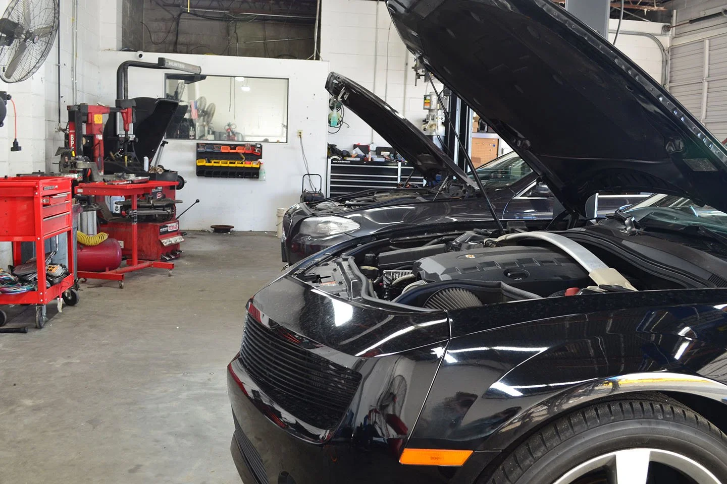A black sports car with its hood open inside a car repair garage, with another black car partially visible in the background. The garage has tools and equipment on red rolling carts and a white wall with a window and storage shelves.