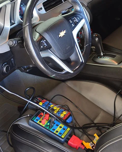 Close-up of a Chevrolet vehicle's interior showing the steering wheel, gear shift, and a device with multiple colorful plugs and wires connected to it.