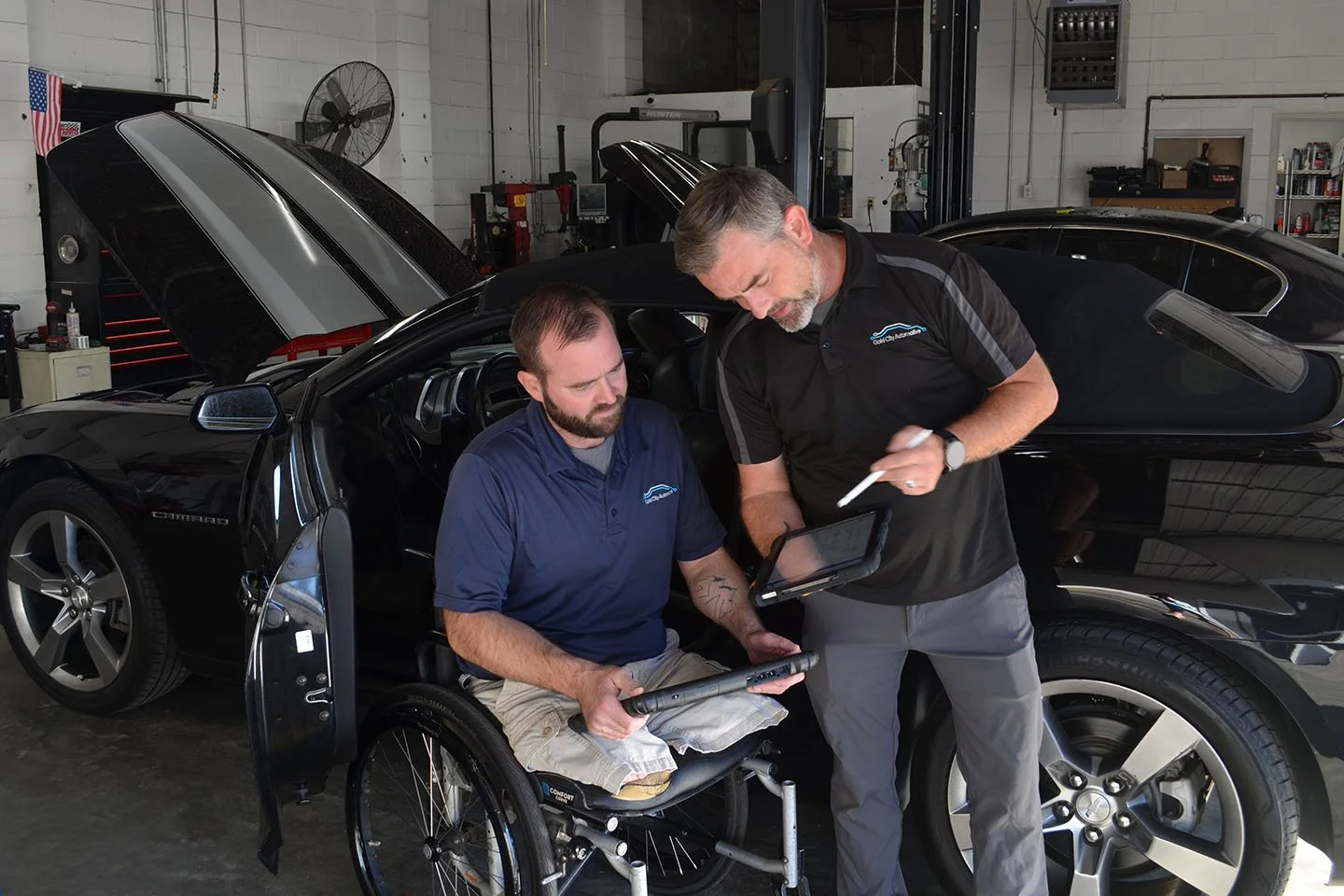 A man in a wheelchair and a man in work attire are inside an auto repair shop, discussing a black sports car. The man in the wheelchair is holding a tablet and looking at the car, while the other man is pointing at a device, possibly a diagnostic tool, and the car has its hood open.