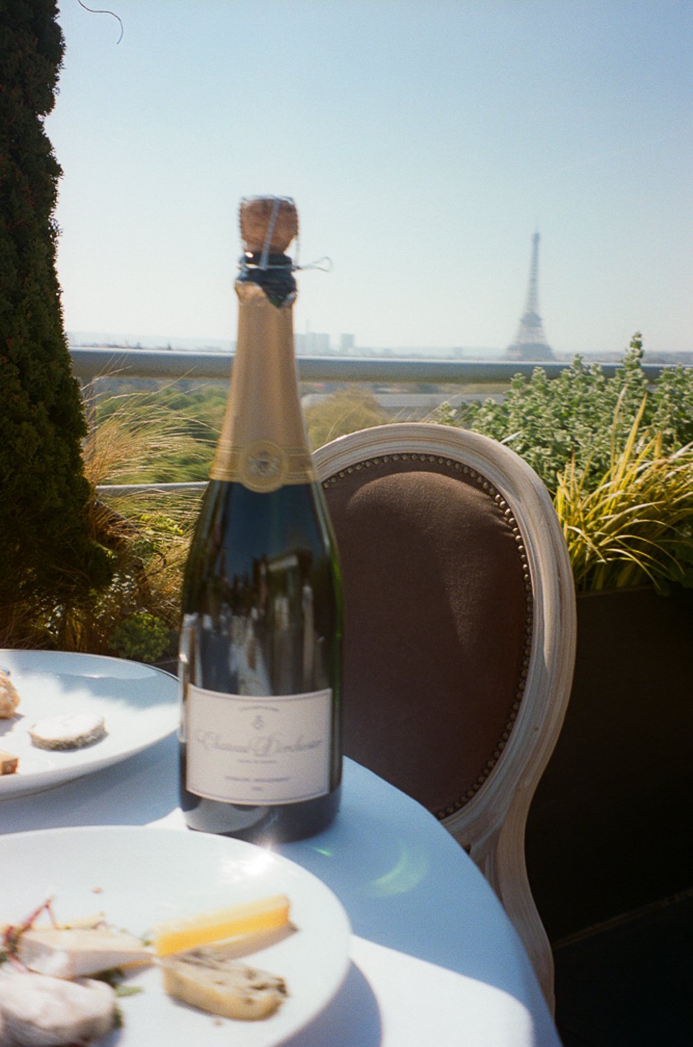 A table set with plates of food, a bottle of champagne, and a chair with a view of the Eiffel Tower in the background on a sunny day.